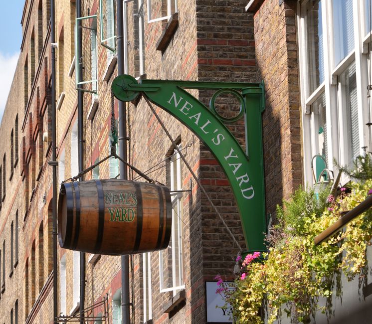A close-up view of a green metal sign reading 'Neal's Yard' attached to a brick building in Covent Garden, London, with a wooden barrel-shaped flower display hanging beneath it. The barrel is marked w