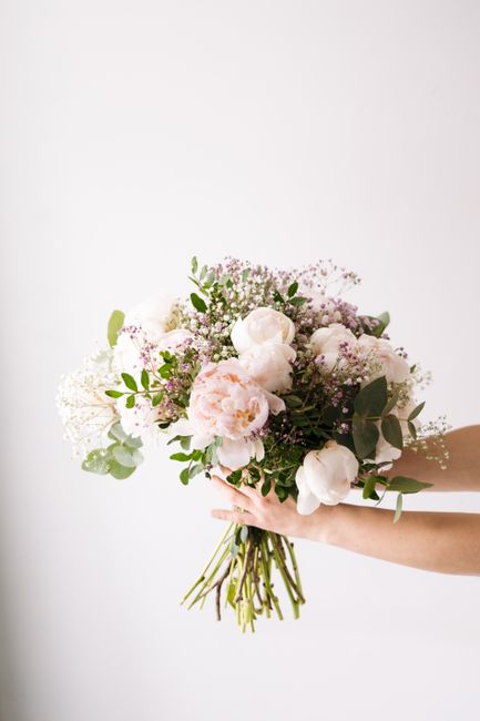 A bouquet of fresh, pastel-colored flowers held by a person's hands against a plain white background. The arrangement features large, soft pink and white peonies, complemented by smaller white blossom