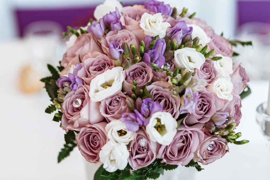 A round floral bouquet featuring light pink roses, white lisianthus, and purple freesia, arranged with green foliage. The flowers are fresh, with delicate petals and sturdy stems, and the bouquet is s