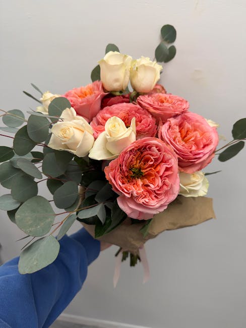 A hand holding a bouquet of fresh flowers against a plain light background, featuring large pink ranunculus flowers with ruffled petals, cream and pale pink roses with smooth, delicate petals, and spr