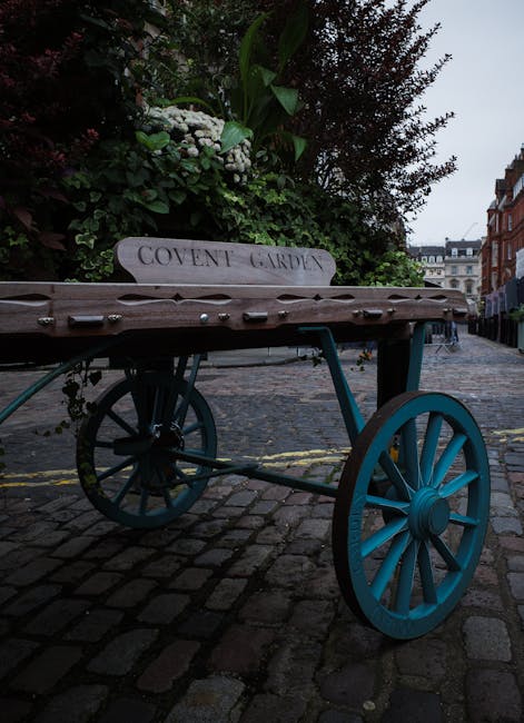 A vintage wooden flower cart with turquoise-painted wheels, placed on a cobblestone street in Covent Garden. The cart features a white wooden sign with the inscription 'COVENT GARDEN' and is topped wi
