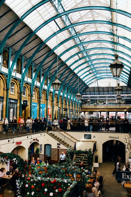 The image depicts an indoor market with a high, arched glass and metal ceiling supported by teal-painted beams. Below, there are multiple levels with a balcony overlooking the main space, populated by