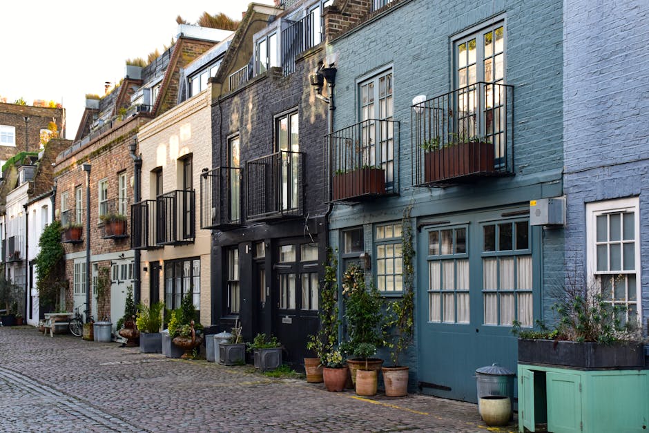 A row of charming, narrow terraced houses with colorful facades, including shades of blue, black, beige, and white, in a cobblestone street. Each house features large front windows, some with small ba