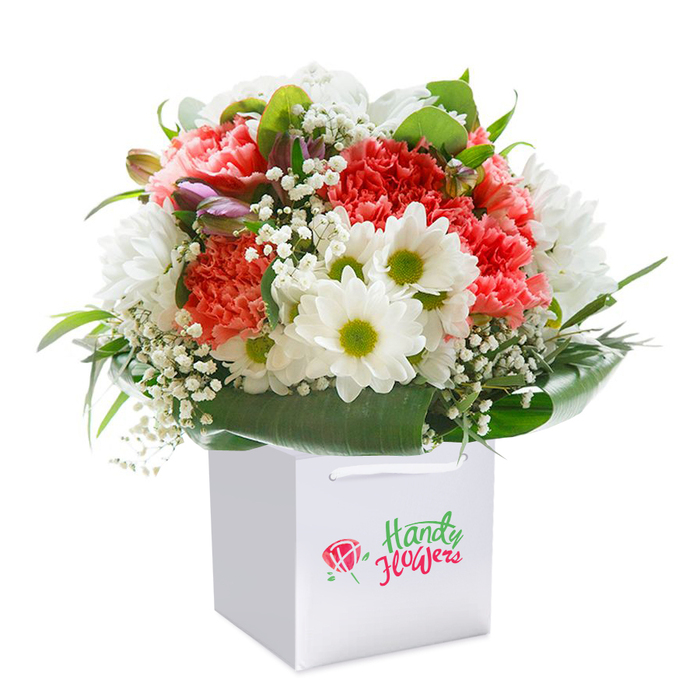 Perfectly poised in a spotless square white gift bag, this radiant Covent Garden bouquet is a testament to local craftsmanship, its gathered blooms evoking the lively spirit of the neighbourhood's flower stalls. The star attractions-volumes of coral and peach carnations-display layers of delicately fringed petals, their soft hues drawing the eye with gentle allure. Bright, white daisies pepper the design, each flower ringed by snowy petals and crowned with expressive yellow-lime centres, imparting a sense of optimism and joy reminiscent of early spring weekends by the Apple Market. Finely textured gypsophila is artfully scattered throughout, lending a dewy, airy softness that sets off the bolder blossoms. Discreet, purple-tinged buds peek from the edges, their unopened faces hinting at new beginnings and giving the bouquet added dimension. All elements are anchored by sweeping arcs of glossy, deep green foliage, carefully framing the arrangement much like the leafy avenues tucked behind Covent Garden's bustling streets. The bouquet forms a sumptuous, rounded dome, radiating freshness and attention to detail. Still-life clarity is achieved with bright, even lighting and a seamless white background, allowing every colour and curve to stand distinct. This engaging composition, ready for delivery or a walk home through Maiden Lane, speaks to moments worth celebrating-birthdays, anniversaries, or simple acts of kindness-making it a cherished expression of thoughtfulness for anyone in Covent Garden.