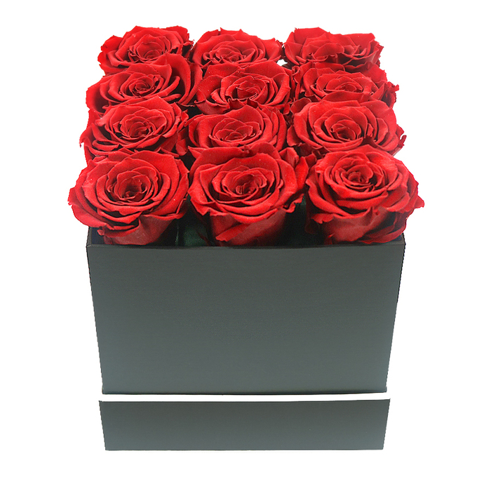 This detailed image presents a lavish arrangement of twelve red roses, meticulously placed in a square black box that seems perfectly at home in the refined, creative atmosphere around Covent Garden. Captured from a straight-on, eye-level perspective, the viewer sees the roses filling the entire mouth of the container, their circular blooms lined up in three precise rows of four. Each rose is in full blossom, its petals unfurling in tightly layered whorls that create a sense of old-world romance paired with modern design. The petals display a luscious, deep crimson tone, almost jewel-like, with slightly darker shadows tucked into the centre and softer, brighter edges that catch the light.
The box below is a study in minimalist elegance: a square, matte black form with hard, clean lines and a smooth, non-reflective surface. Around its lower portion runs a narrow white band, neat and uninterrupted, adding a tailored detail that evokes the crisp signage and contemporary interiors of the small boutiques tucked between Bow Street and the Covent Garden Piazza. Against the pristine white background, there are no distractions-only the interplay of rich red, stark black, and bright white.
Gentle, even lighting falls from above, softening the shadows while still defining the sculpted shapes of petals and box. The preserved roses sit proudly just above the rim, suggesting longevity and permanence, a fitting choice for long-lasting romantic gifts sent to homes, hotels, or offices across WC2. The overall composition feels poised and considered, evoking feelings of enduring affection, special anniversaries, Valentine's surprises, and the timeless gesture of giving red roses in one of London's most iconic neighbourhoods.