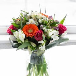 This opulent Covent Garden bouquet, artfully displayed in a slender glass vase, is a testament to the local florists' attention to detail and love for their craft. The central stars are bold orange gerbera daisies and elegant peach-to-cream varieties, their large petals layered like soft rosettes around deep, chocolate-hued centres. Set beside them, plush magenta roses unfurl with a sense of refined drama, invoking the romance and energy of Covent Garden's theatre district. Interspersed among these are airy clusters of white alstroemeria and petite spray blossoms, which lend a subtle radiance and gentle texture to the composition. Under the soft, ambient light that filters in, every colour glows - from the delicate blush-pink highlights to the crisp white accents that offset deeper tones. Broad, glossy aspidistra leaves artfully embrace the lower half of the bouquet, wrapping it with a verdant collar reminiscent of the greenery in nearby Neal's Yard. Smaller pointed leaves and feathery sprigs weave between each bloom, giving the whole arrangement a layered, garden-fresh feeling. The stems, revealed through the vase's clarity, add a further note of authenticity and freshness. There is a sense of flowing movement and joyful abundance, heightened by the gentle mingling fragrance of rose and lily. A bouquet like this would perfectly grace a local brunch on a sun-dappled Covent Garden morning or offer a warm gesture to mark new beginnings, its optimism and elegance echoing the vibrancy of this beloved London area.