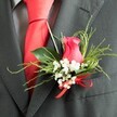 The image offers an intimate close-up of a wedding boutonniere fastened to a charcoal grey suit jacket, evoking the polished elegance of a ceremony near Covent Garden's cobbled streets. The centrepiece is a single deep pink rosebud, tipped toward fuchsia, its petals tightly layered yet beginning to unfurl, creating a sense of anticipation and quiet romance. The rose is encircled by a delicate ring of white baby's breath, each miniature blossom crisp and bright, standing out clearly against both the dark wool of the lapel and the saturated pink of the central bloom. Surrounding greenery in varied textures brings balance: broad, shiny leaves sit snugly against the jacket, while slimmer, feathery fronds arc outward, adding lightness and a gentle sense of motion. At the base of the flowers, a slim red satin ribbon is tied into a neat, symmetrical bow, its vivid scarlet hue mirroring the smooth, plain red silk tie tucked beneath the lapel. The tie lies cleanly over a subtly patterned grey dress shirt, whose discrete design softens the strong colour contrast without drawing focus away from the floral detail. The fabric of the suit appears refined and slightly textured, suggesting a fine wool blend. Overall, the boutonniere feels carefully composed and confidently styled, the kind of detail lovingly prepared for a groom or groomsman attending a wedding or formal event in Covent Garden. It suggests a moment of poised calm before the vows, where a small cluster of flowers adds meaning, colour, and personal expression to a sharply tailored ensemble.